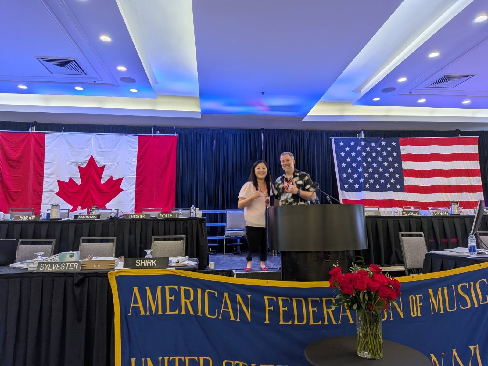 Rebecca Matayoshi and John LeBlanc at the podium between the Canadian and US flags